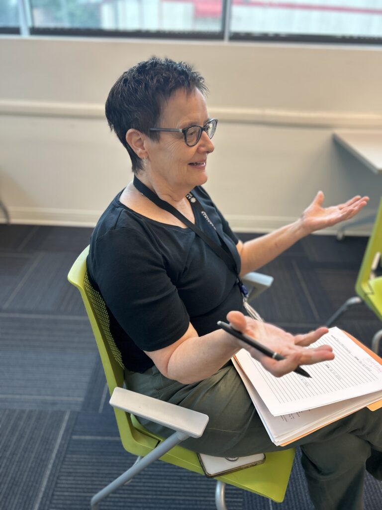 A person wearing a black shirt and green pants with a notebook in their lap talking to people.
