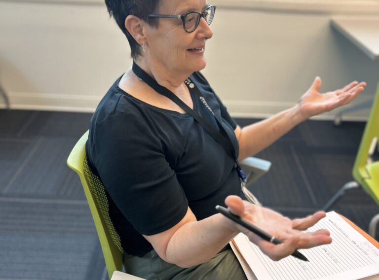 A person wearing a black shirt and green pants with a notebook in their lap talking to people.
