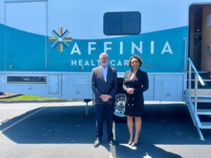 A man in a dark suit standing next to a woman in a dark suit in front of a bus labeled Affinia Healthcare.