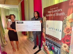 A Black woman in a black dress and a Black man in a gray suit are holding a check in a decorated hall with pink and green drapes and a sign that says The Clayco's Garden of Hearts.